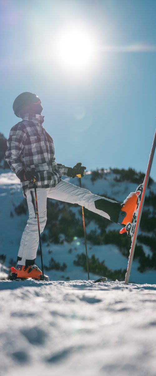 A mid-adult Asian male takes a break from skiing, standing with equipment on a snowy mountain slope under a clear blue sky, wearing winter sportswear and protective gear.